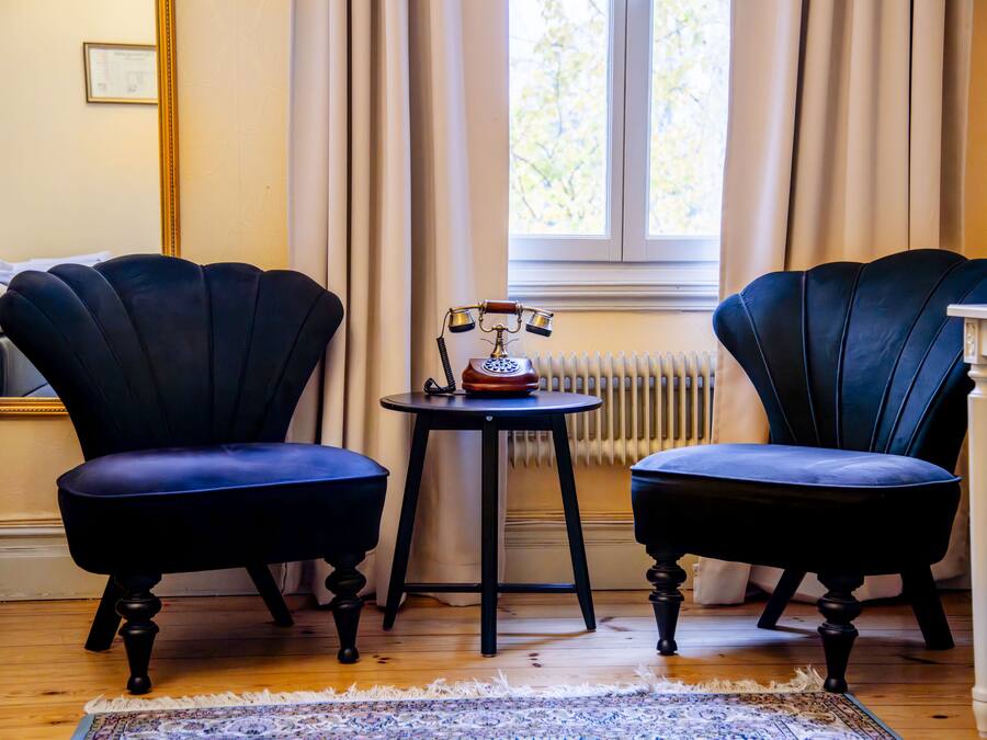 Una habitación con dos sillones tapizados en azul oscuro, una mesita redonda con un teléfono antiguo, un radiador y una ventana con cortinas.