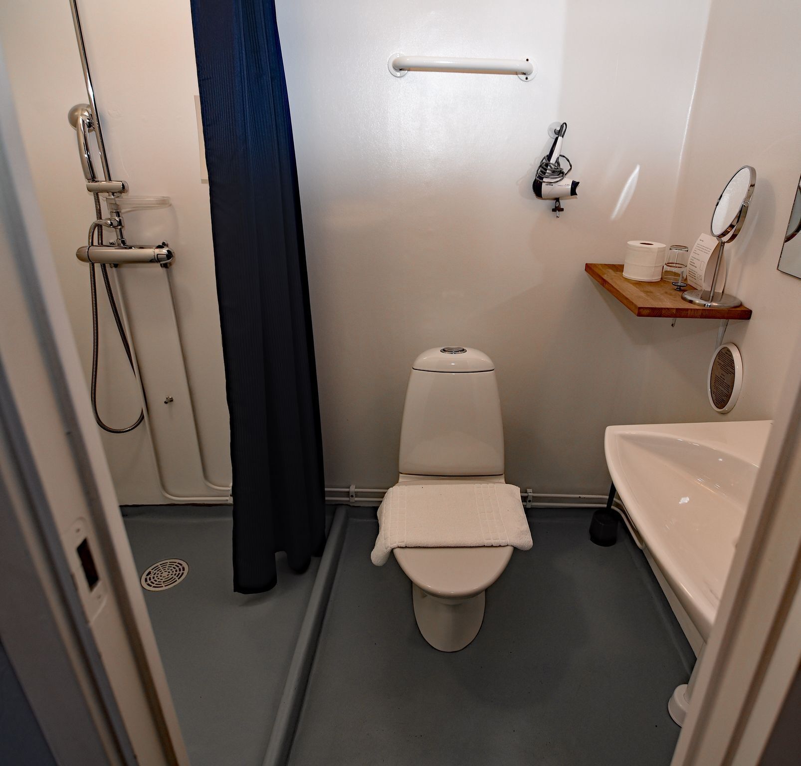 A compact bathroom with a toilet, a shower, a sink, and a small shelf with toiletries.