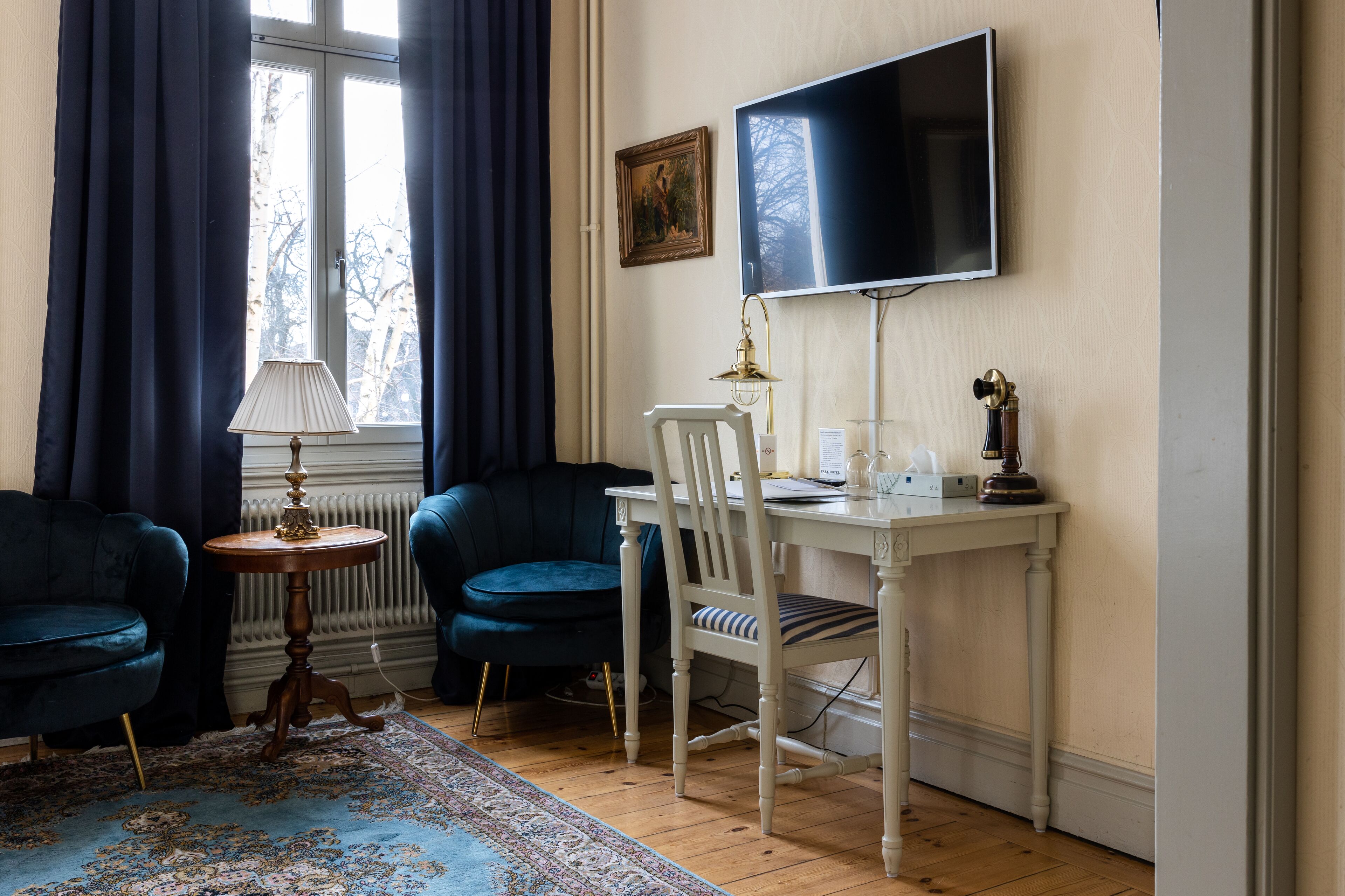 Una habitación con un escritorio blanco, un sillón azul, otro sillón oscuro, una pequeña mesa redonda con una lámpara, y un televisor grande montado en la pared.