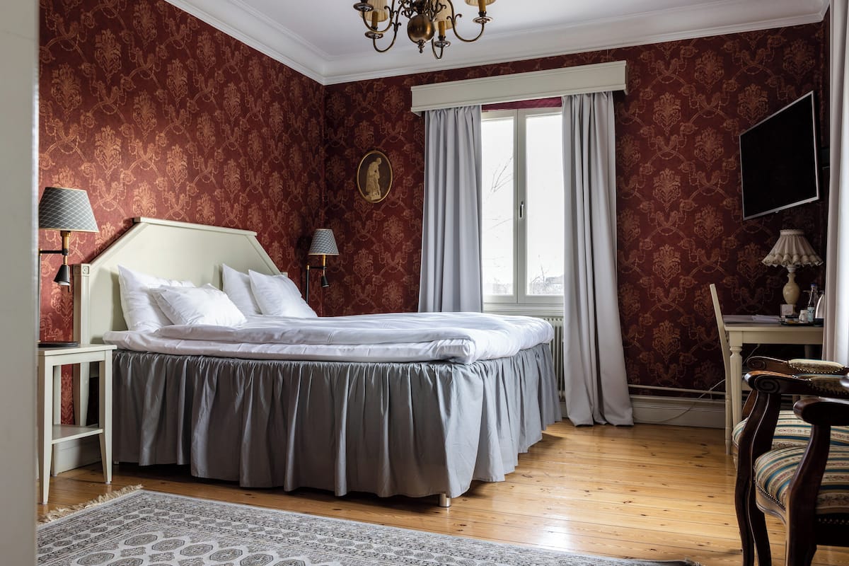 Un dormitorio con cama, escritorio, silla, ventana con cortinas y candelabro.