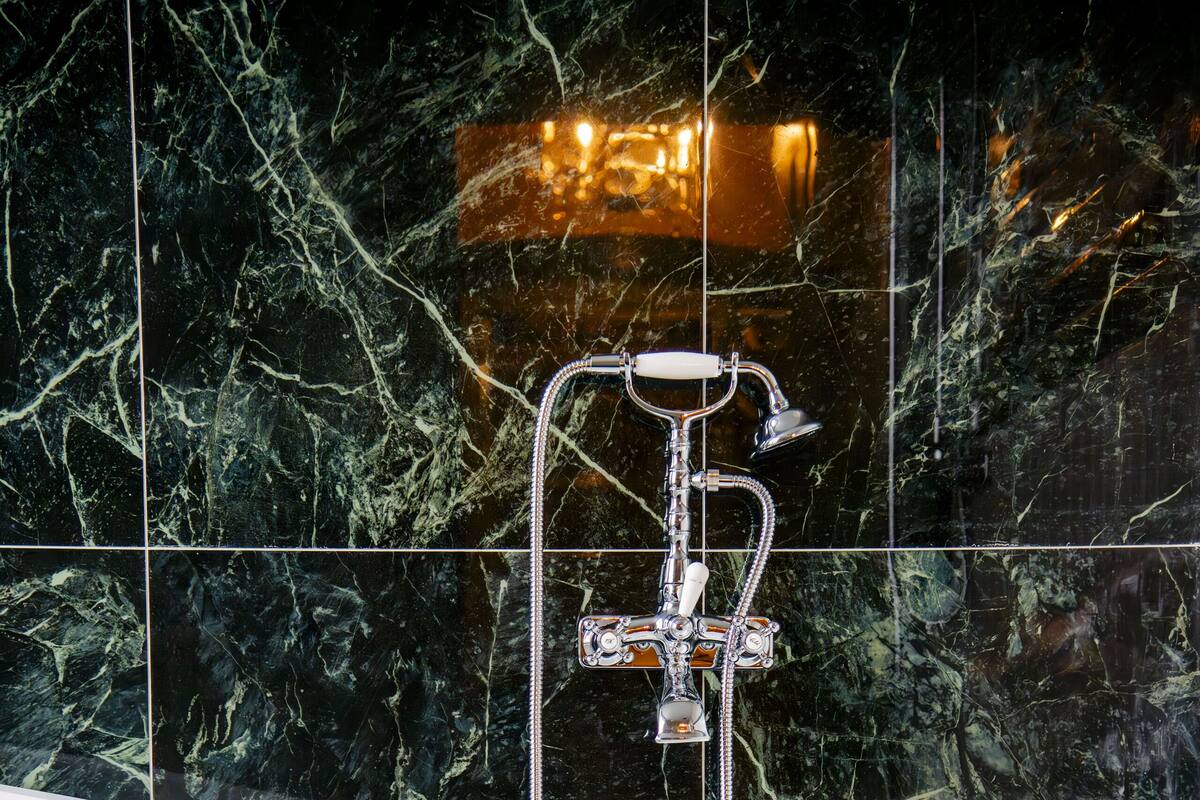 Una bañera independiente con grifo y ducha de cromo, frente a una pared de mármol oscuro.