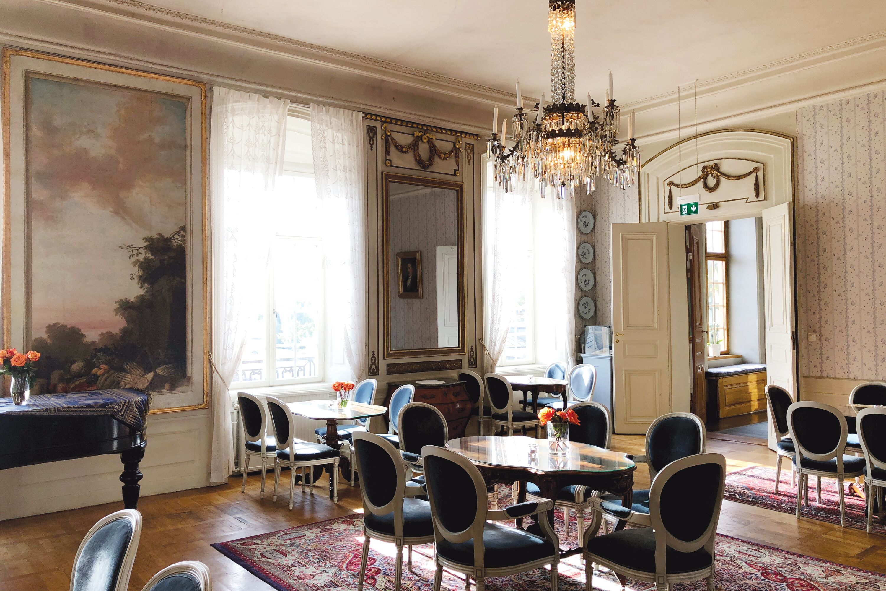 A dining area with multiple tables and chairs, a chandelier, and a large painting on the wall.
