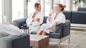 Deux personnes vêtues de longues robes blanches sont assises sur un canapé gris dans une pièce bien éclairée, dotée de grandes fenêtres.