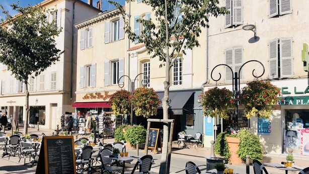 Um café ao ar livre com cadeiras e mesas de vime, rodeado por edifícios com persianas e cestos de flores penduradas.
