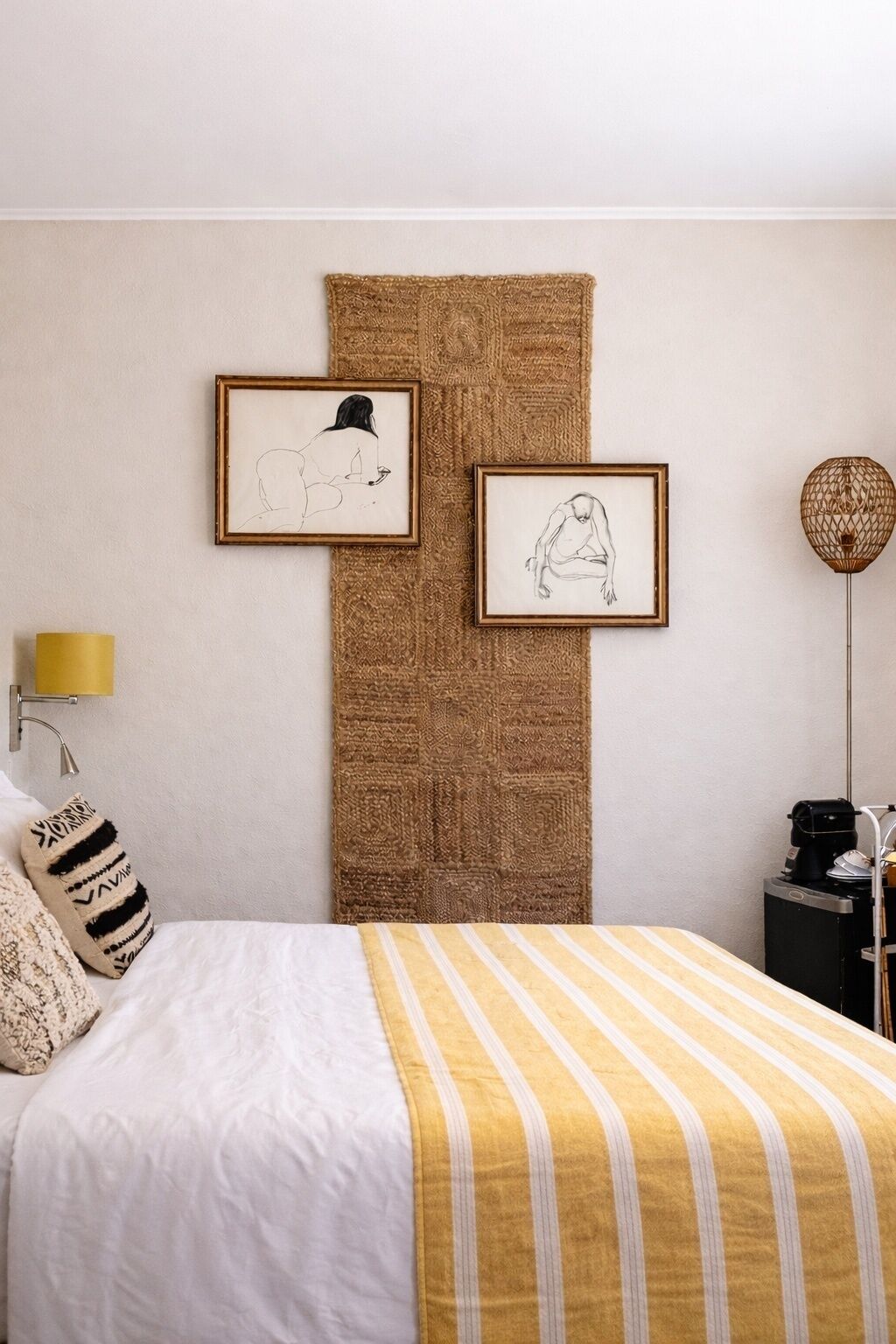 A bedroom with a bed, a wall-mounted lamp, a coffee machine, and two framed artworks.