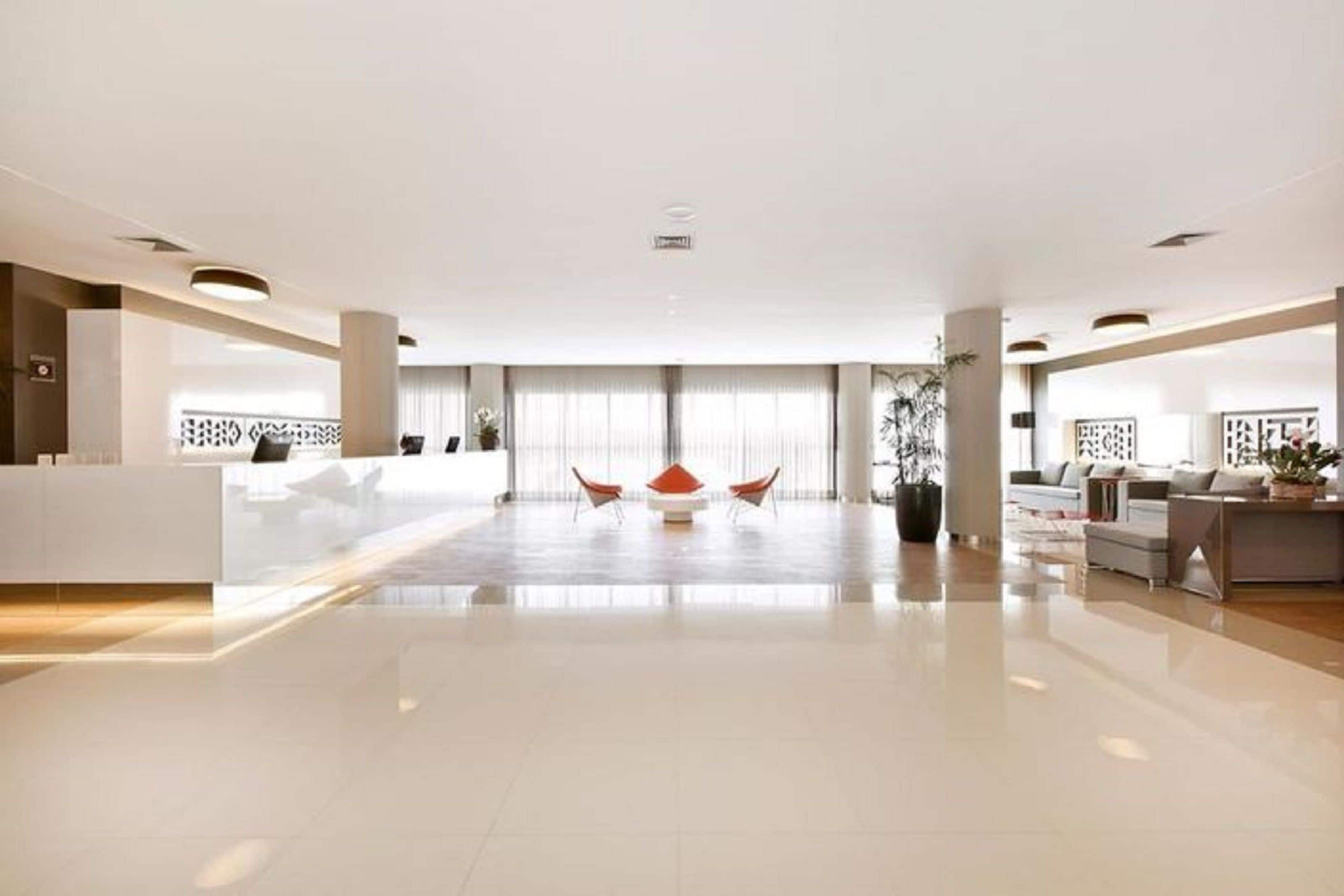 A spacious, modern hotel lobby with a central seating area, a reception desk, and a large window providing natural light.
