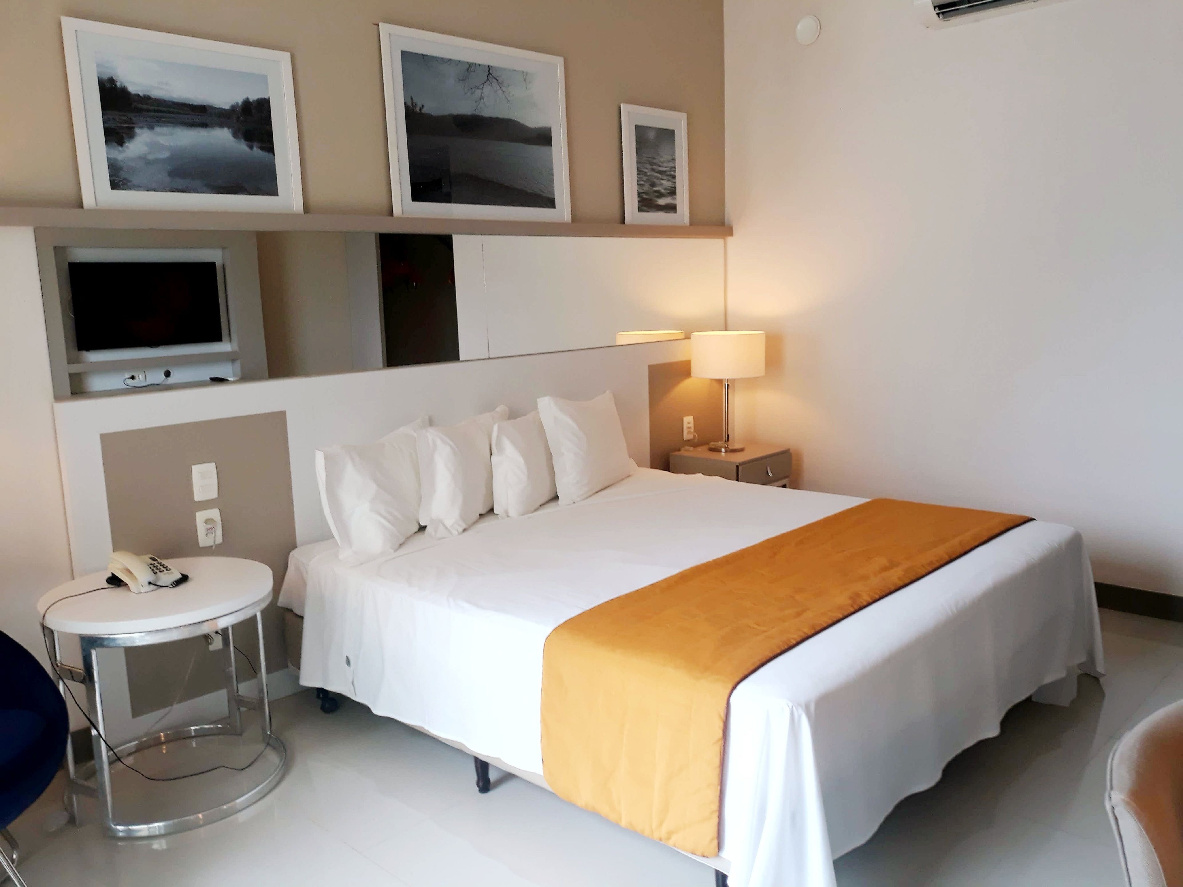 Habitación de hotel con cama, mesita de noche con lámpara, una silla, un escritorio con teléfono y cuadros en la pared.