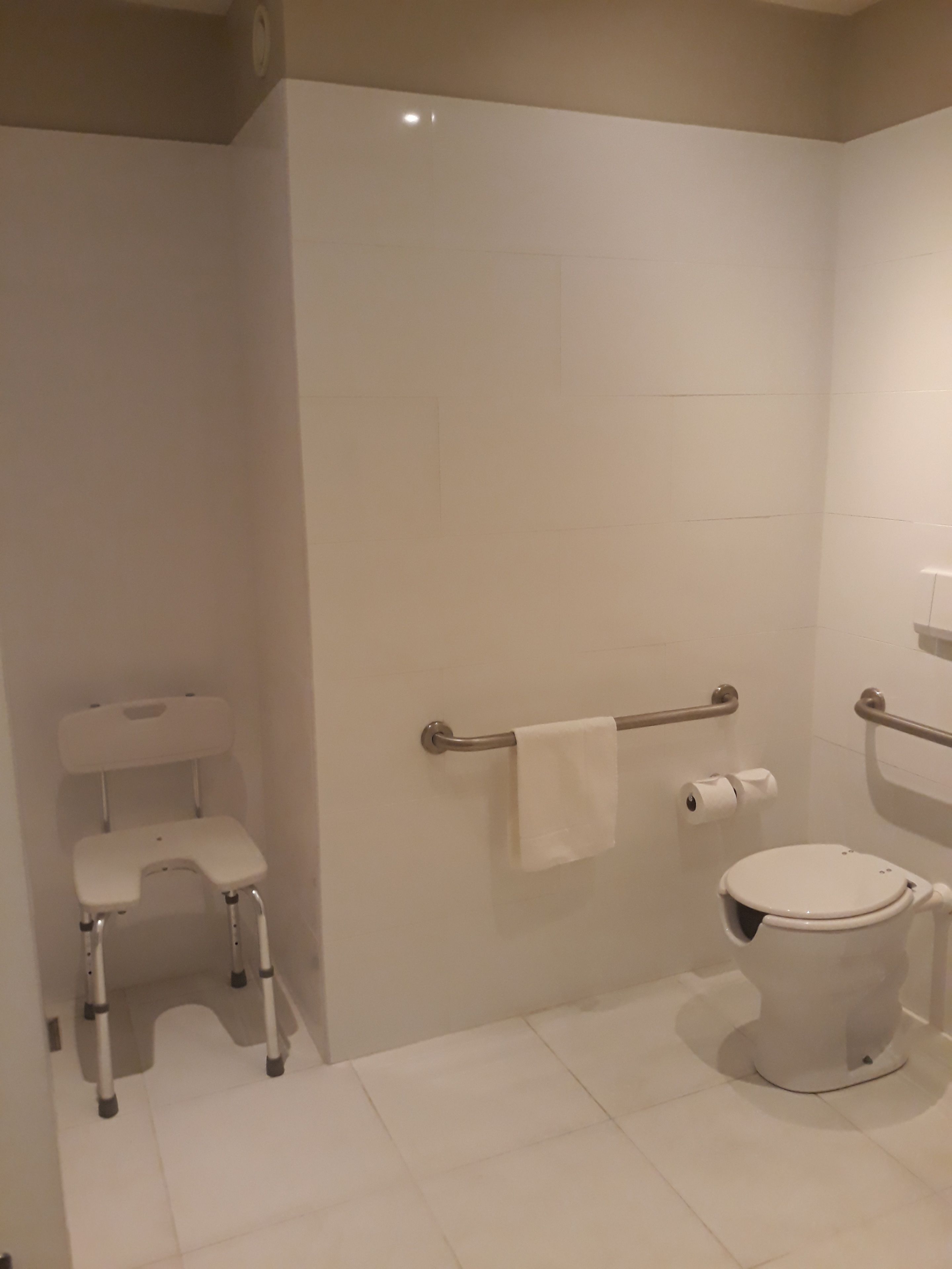 A modern bathroom with a white toilet, a grab bar, a shower chair, and a towel rack.