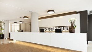 Un área de recepción moderna con un mostrador blanco, dos sillas negras, un jarrón decorativo con flores blancas y una pared con diseño geométrico.