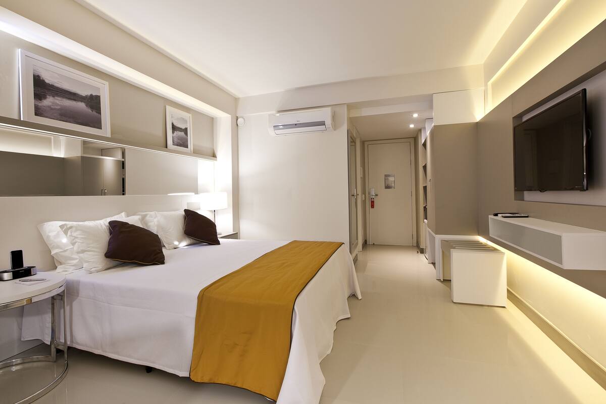 A modern hotel room with a neatly made bed, a bedside table with a lamp, a TV mounted on the wall, and a hallway leading to other rooms.