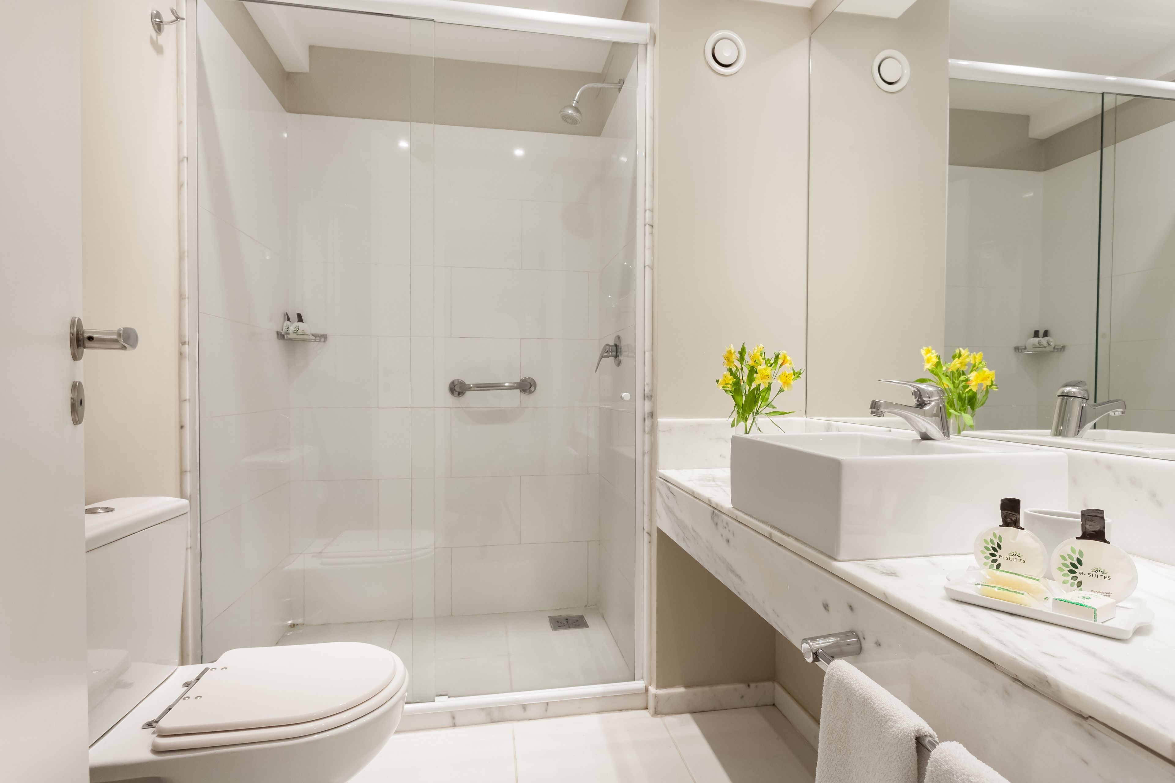 Un baño moderno con ducha de vidrio, inodoro blanco, espejo grande y encimera de mármol con artículos de aseo.