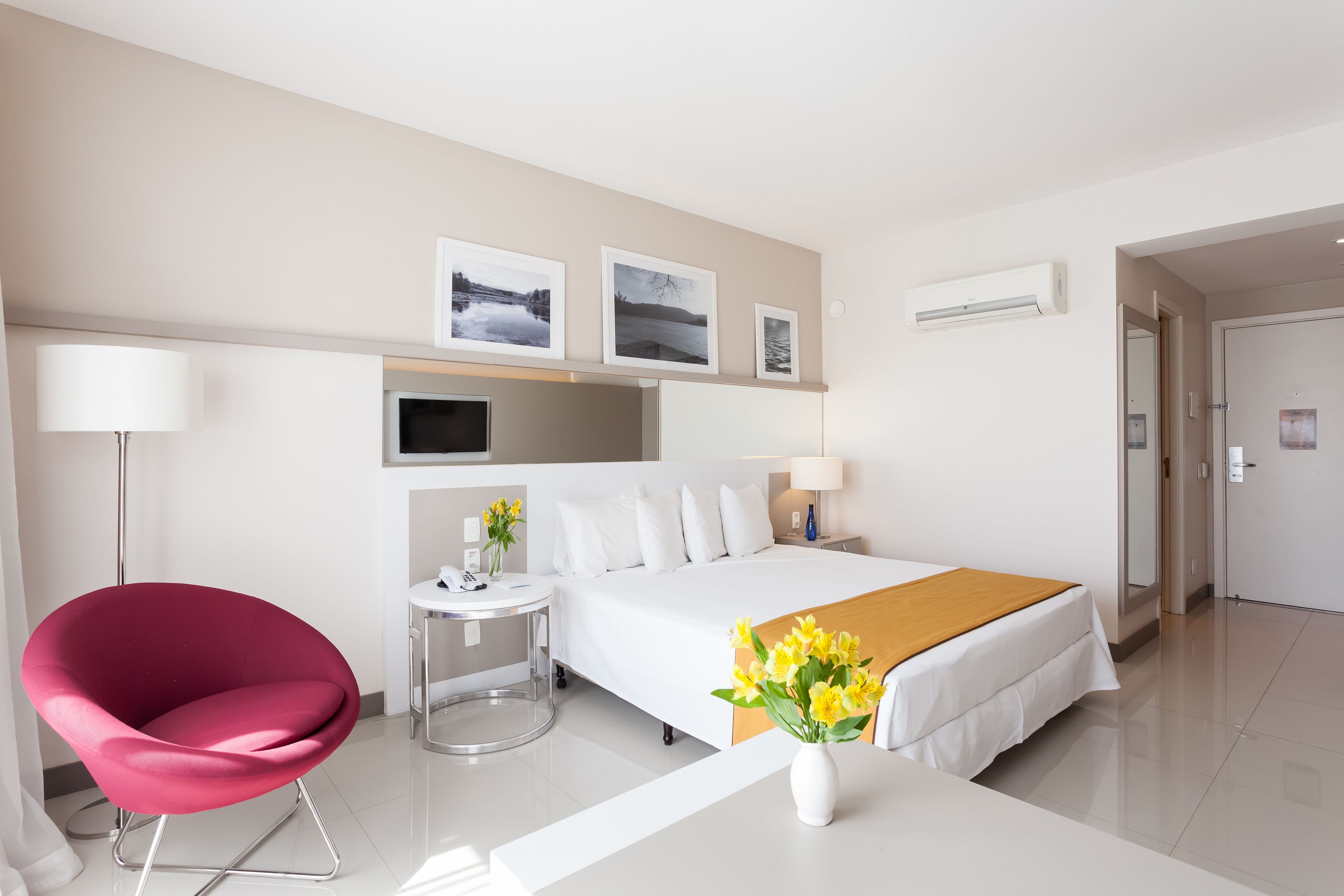 Ein modernes Hotelzimmer mit einem Bett, einem roten Sessel, einem Nachttisch mit Lampe und einem an der Wand montierten Fernseher.