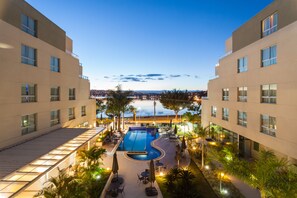 A hotel with a swimming pool, outdoor seating, and a view of the city at dusk.