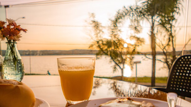 A table with a plate of food and a glass of orange juice, set against a view of a body of water and trees. - Esuites Lagoa Santa (Lagoa Santa)