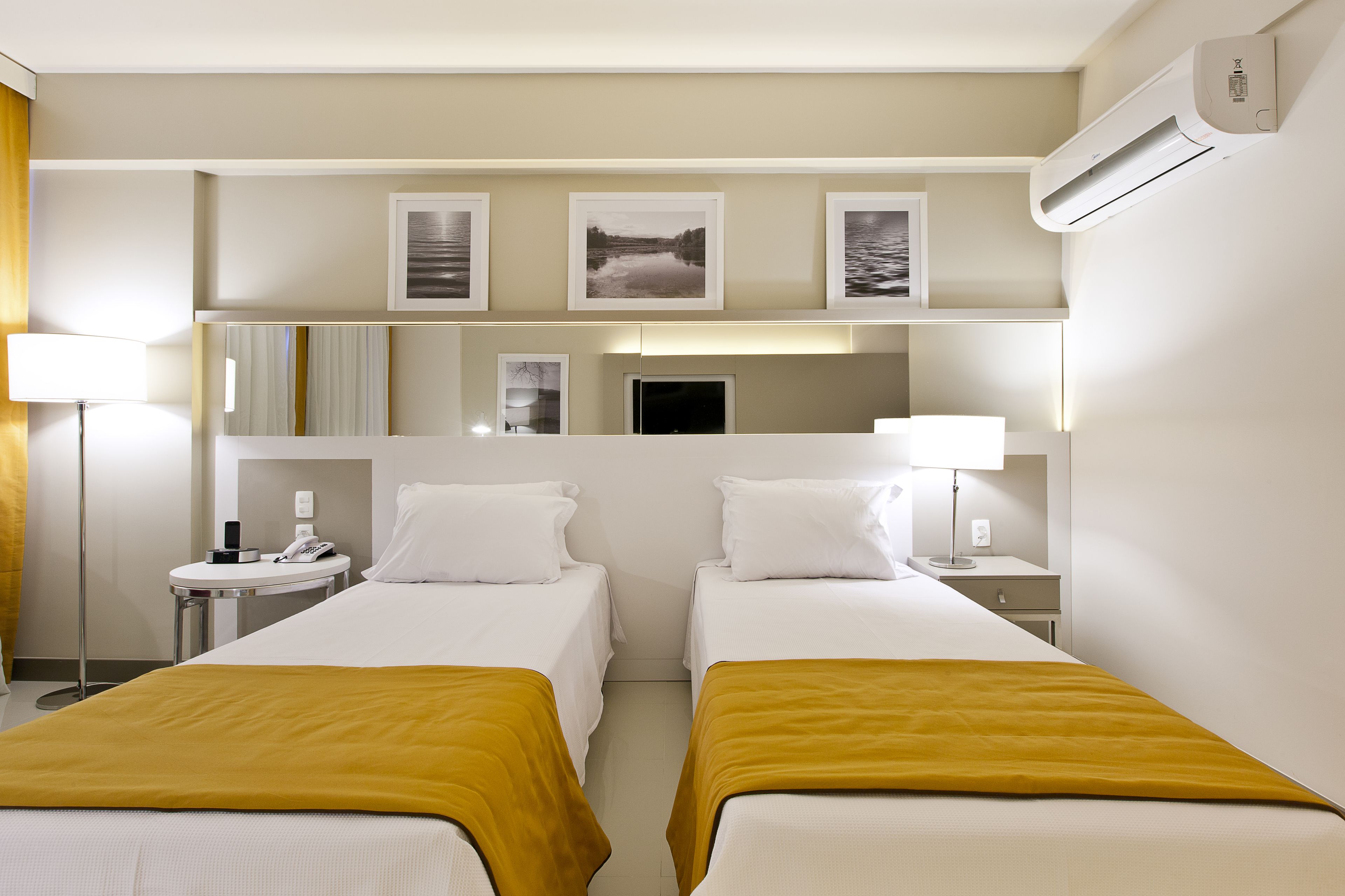 A hotel room with two beds, a bedside table with a lamp, a small round table with a phone, and a wall-mounted air conditioner.