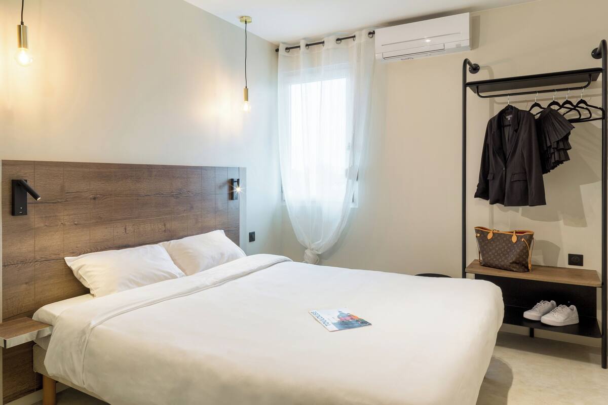 A neatly made bed with a magazine, a wooden headboard, a clothing rack with hangers and a handbag, and a shelf with shoes.