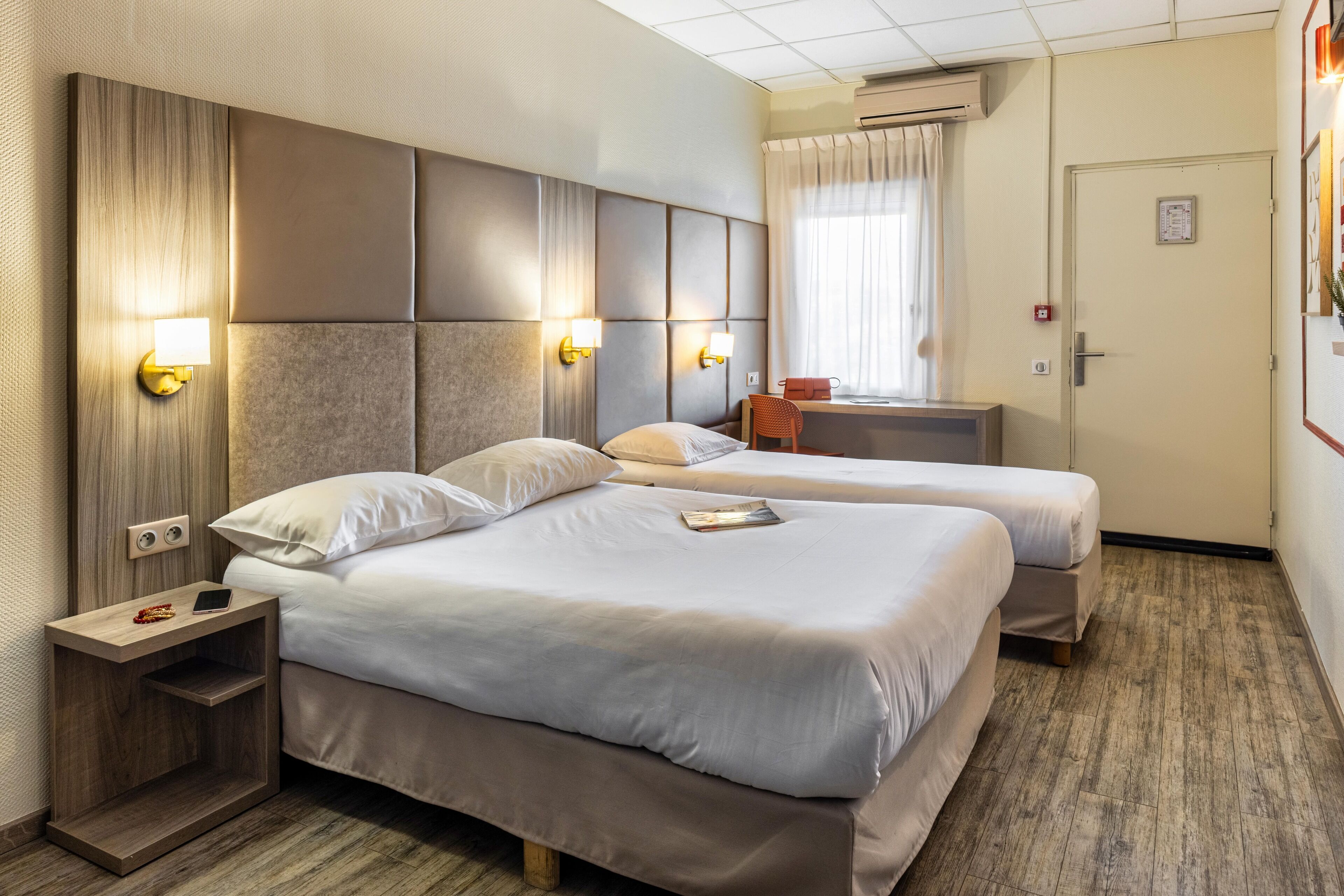 Habitación de hotel con dos camas, un escritorio y una silla. Cuenta con luces instaladas en la pared y un equipo de aire acondicionado.