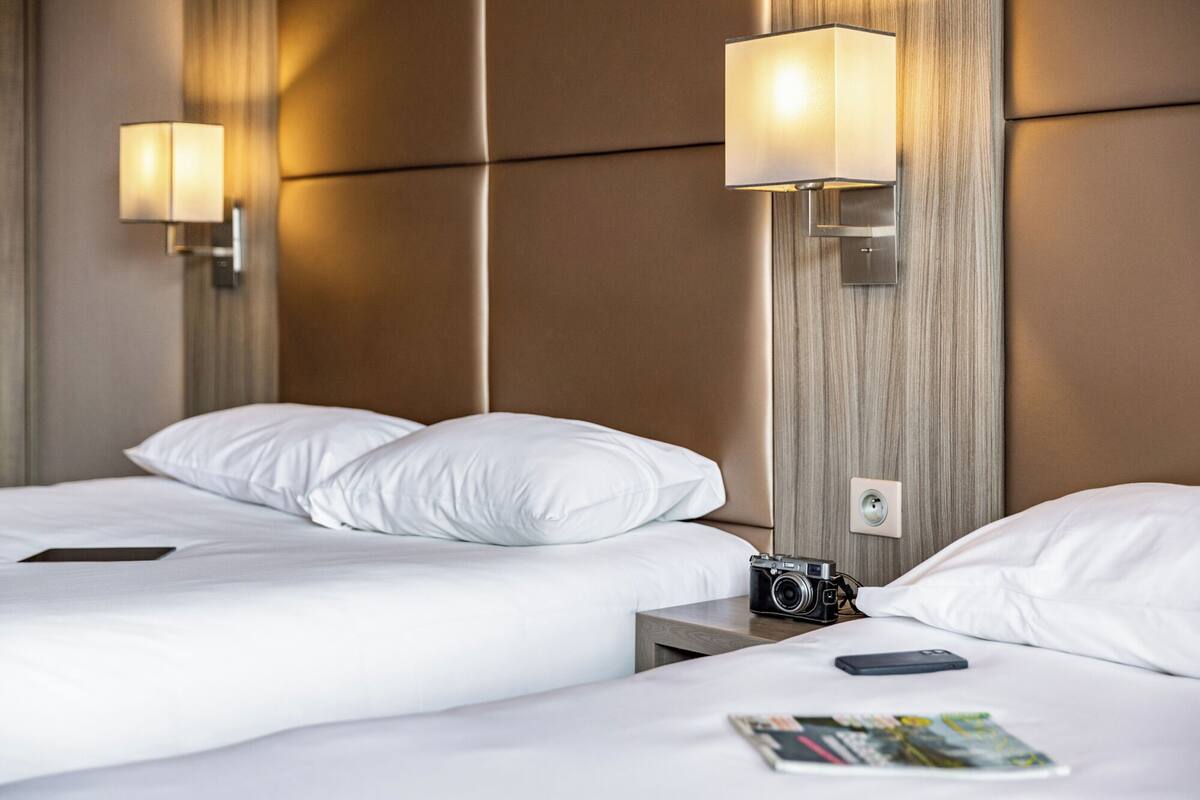 A hotel room with two beds, a nightstand with a camera and a phone, and wall-mounted lamps.