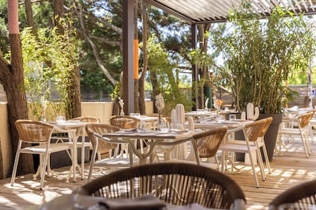 An outdoor dining area with white tables and wicker chairs, surrounded by potted plants and trees.