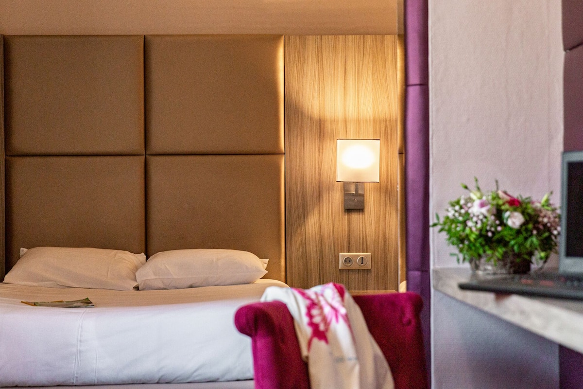A hotel room with a bed, a wooden headboard, a nightstand with a lamp, and a red chair.