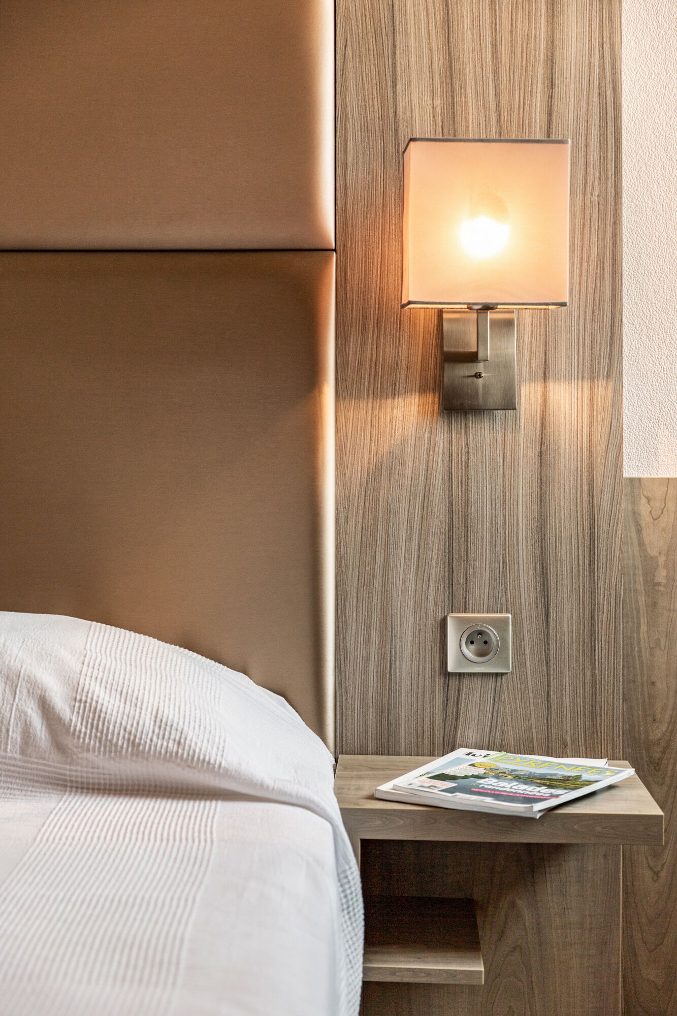 A neatly made bed with a white comforter, a wooden nightstand with a magazine, and a wall-mounted lamp.