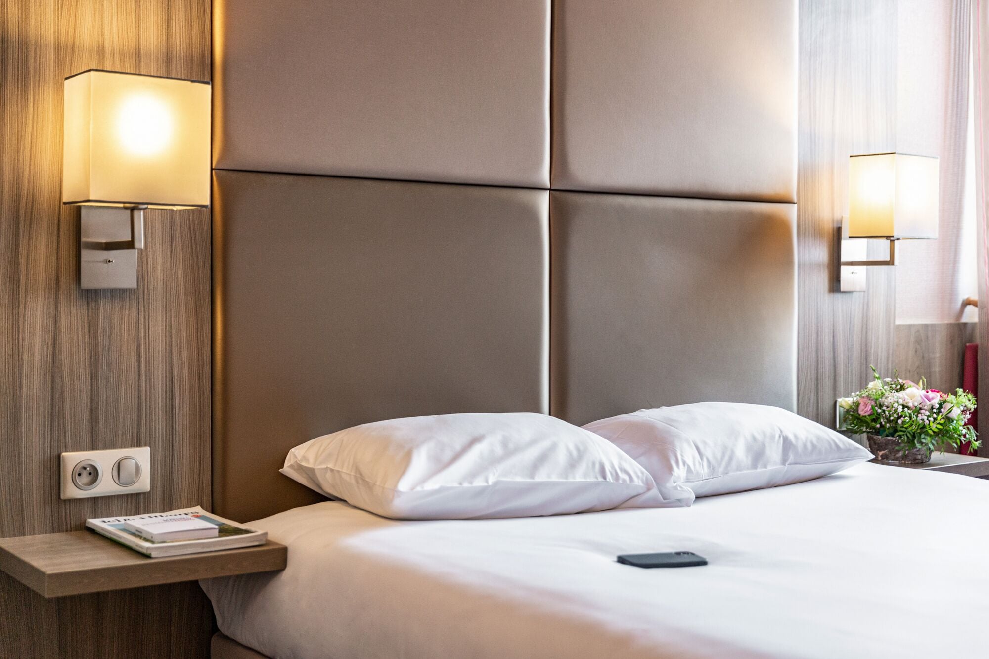Een hotelkamer met een groot bed, twee bedlampen, een nachtkastje met een boek en een stopcontact.