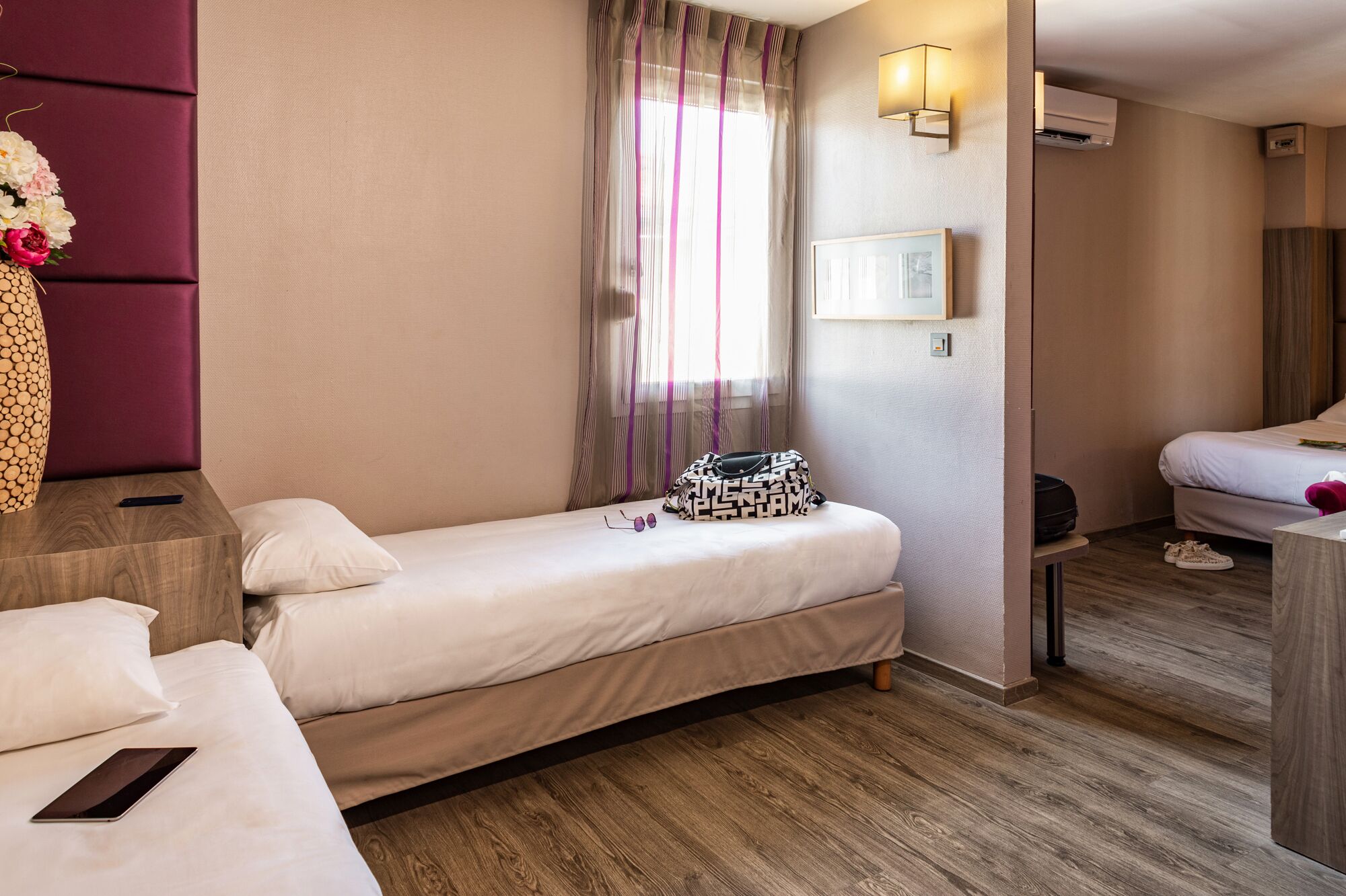 A hotel room with a single bed, a bedside table with a phone, a small table with a handbag, a window with curtains, and an air conditioning unit.
