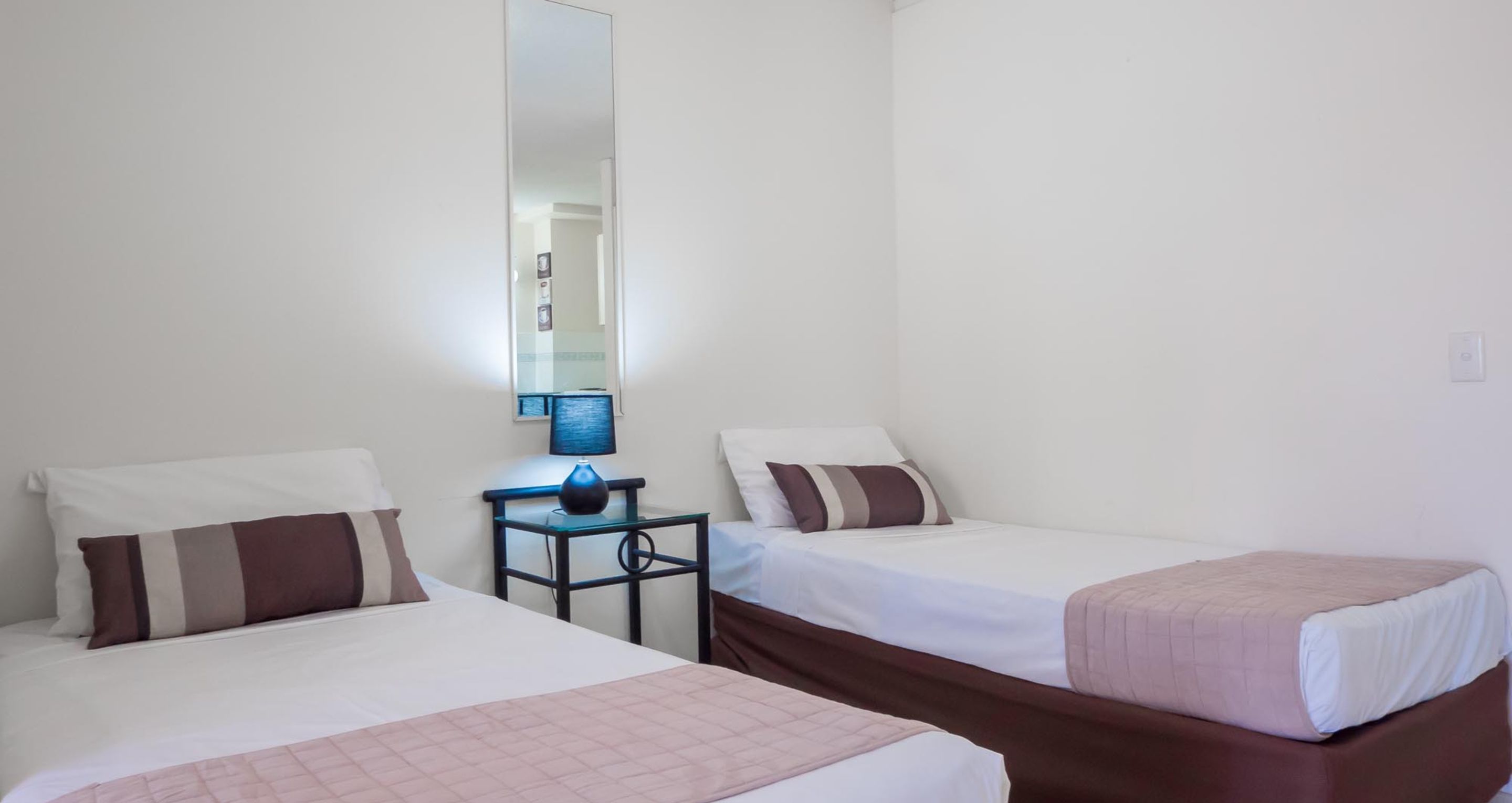 Quarto de hotel com duas camas de solteiro, uma mesinha com abajur e um espelho na parede.