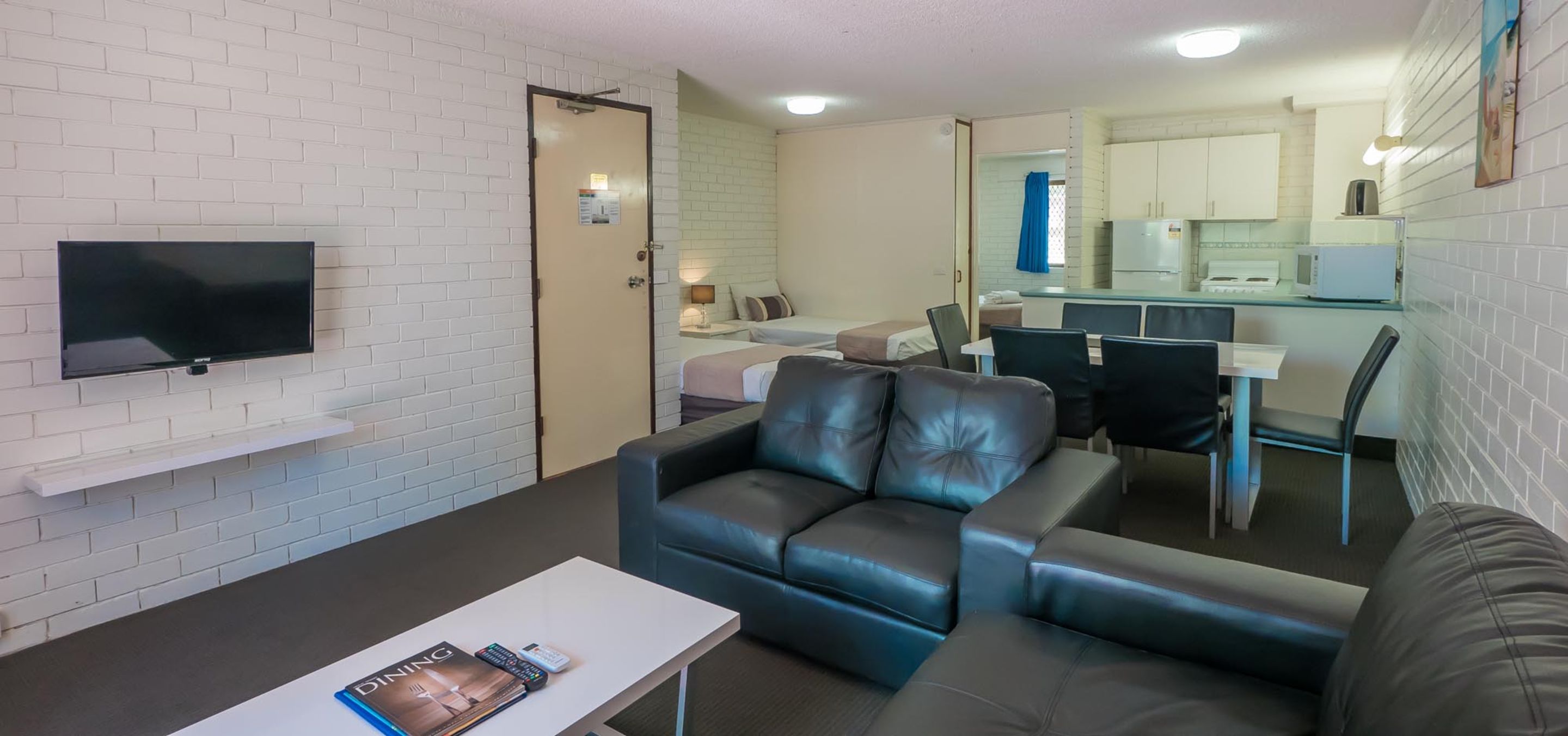 Un espacio habitable compacto con sofá, televisor de pantalla plana, pequeña mesa de comedor y kitchenette al fondo.