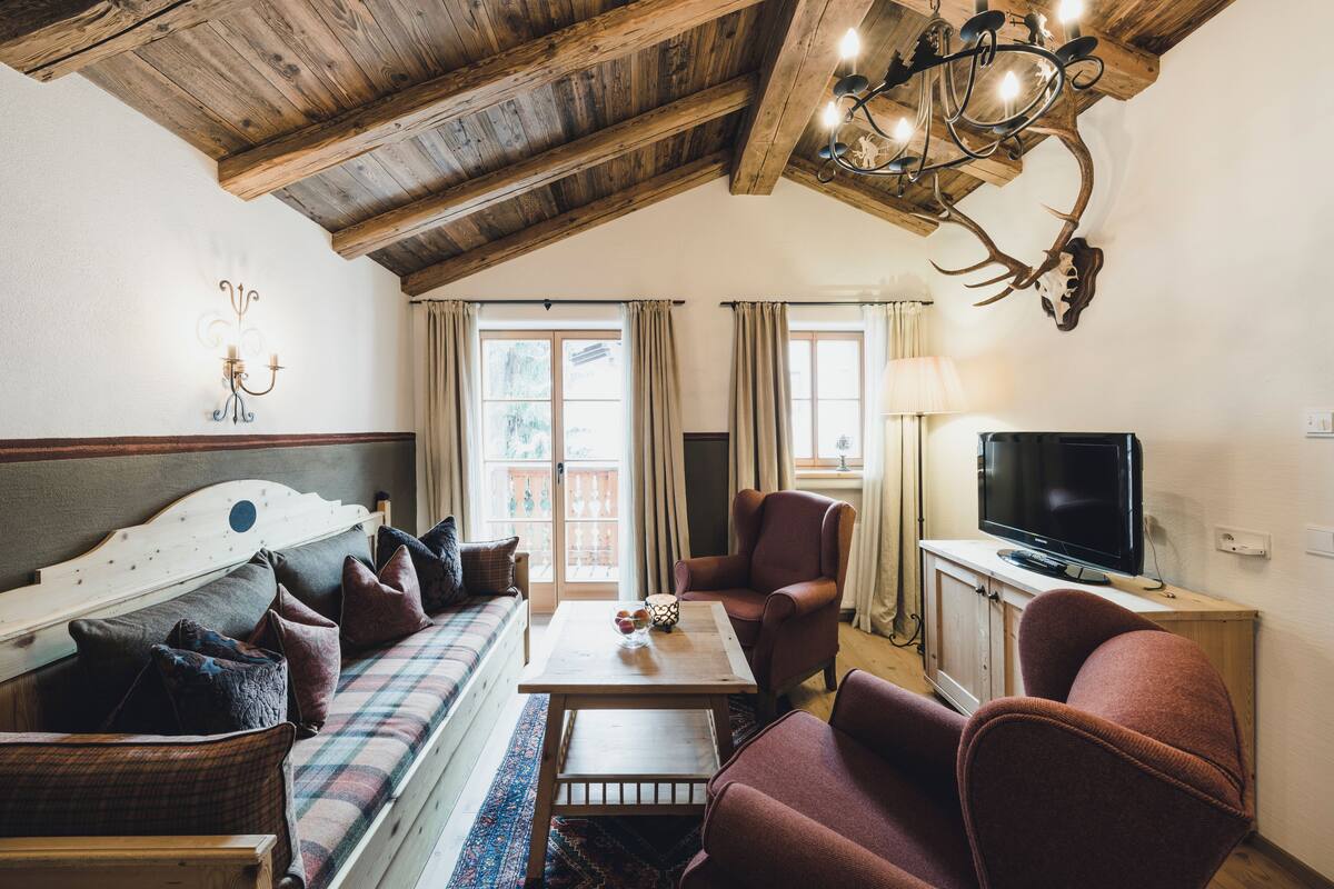 A cozy living room with a wooden ceiling, a sofa, a red armchair, a television on a wooden cabinet, and a chandelier with antlers.
