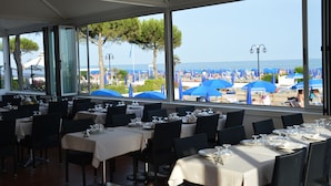 En restaurang med bord dukade för middag och stora fönster som ger utsikt över en strand med parasoller.
