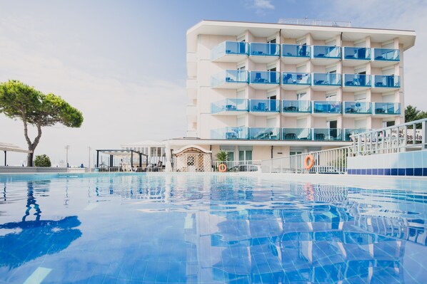 Un hotel moderno con piscina, varios balcones y cielo despejado.