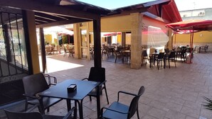 An outdoor patio area with tables and chairs, some covered by red umbrellas, and a fire extinguisher on the wall. - Hotel El Doncel (Atarfe)