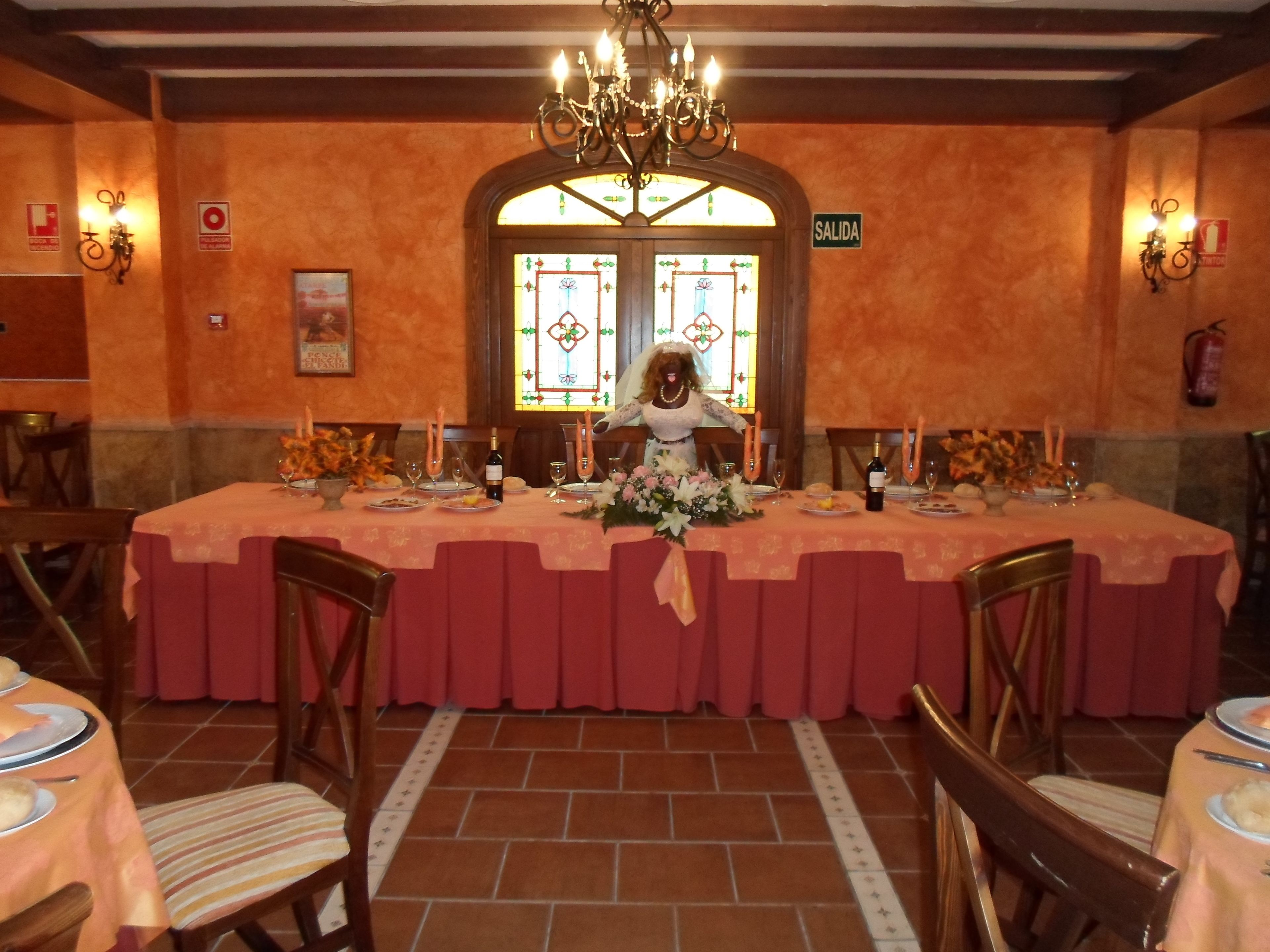 Un comedor con una larga mesa dispuesta para una comida, una araña colgante en el techo y un cartel que indica la salida.