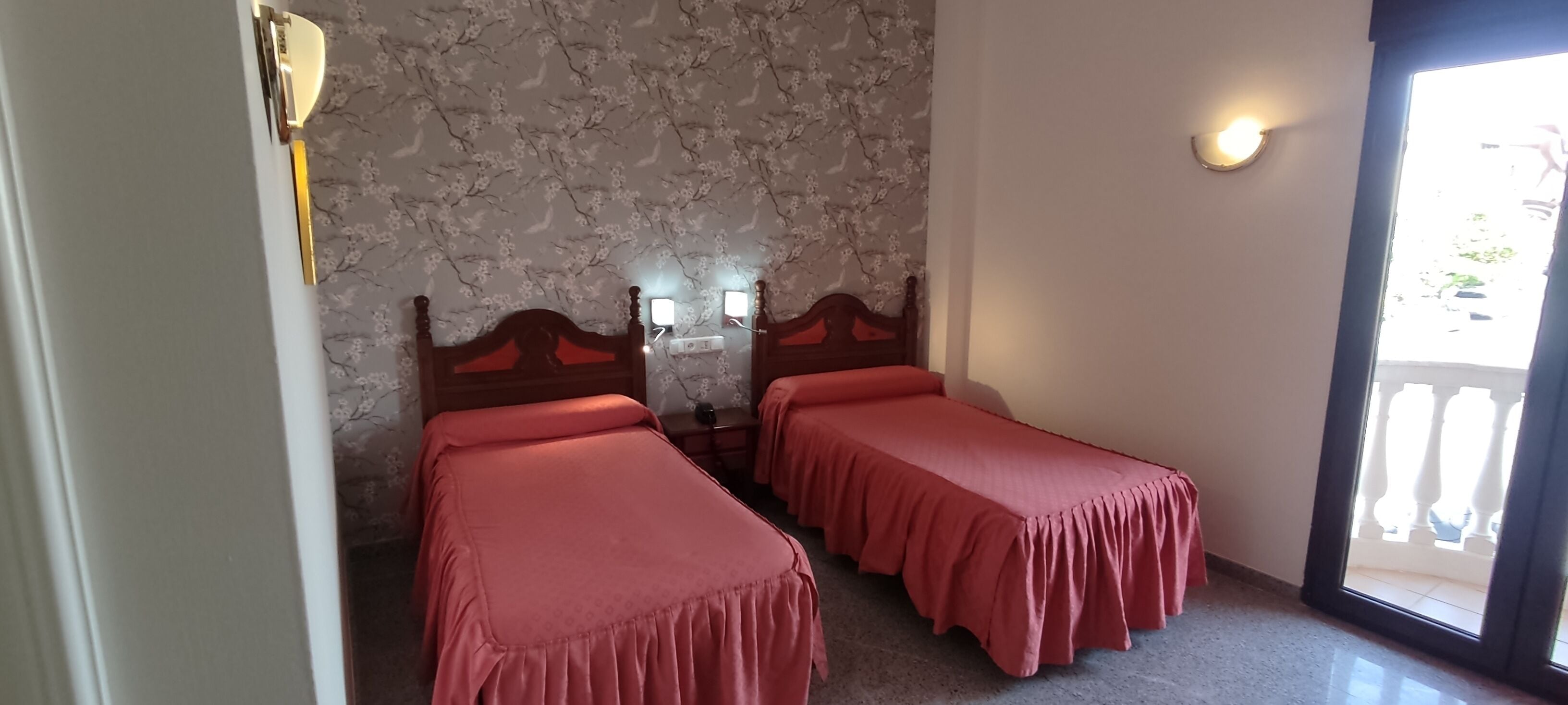 Una habitación con dos camas individuales, cabecero de madera, pared con estampado de piedra, una mesita y una puerta con lámpara.