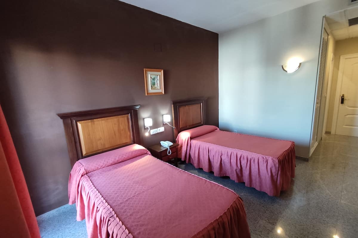 Hotellihuone, jossa on kaksi sänkyä. Jokaisessa sängyssä on puinen pääty ja vaaleanpunaiset vuodevaatteet. Huoneessa on pieni yöpöytä, jossa on puhelin, sekä seinään kiinnitetty valaisin.