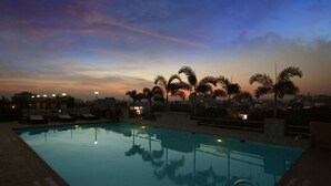 Piscina en la azotea con palmeras y vistas a la ciudad al anochecer.