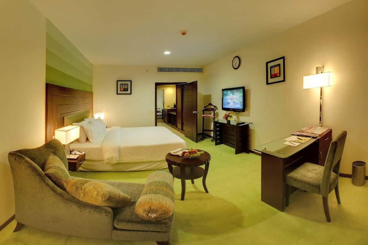 Habitación de hotel con cama, escritorio, silla, una pequeña mesa con fruta, televisor y lámpara.