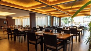 A restaurant with wooden tables and chairs, large windows, and a warm ceiling design. - San Diego Suítes Veredas Sete Lagoas (Sete Lagoas)