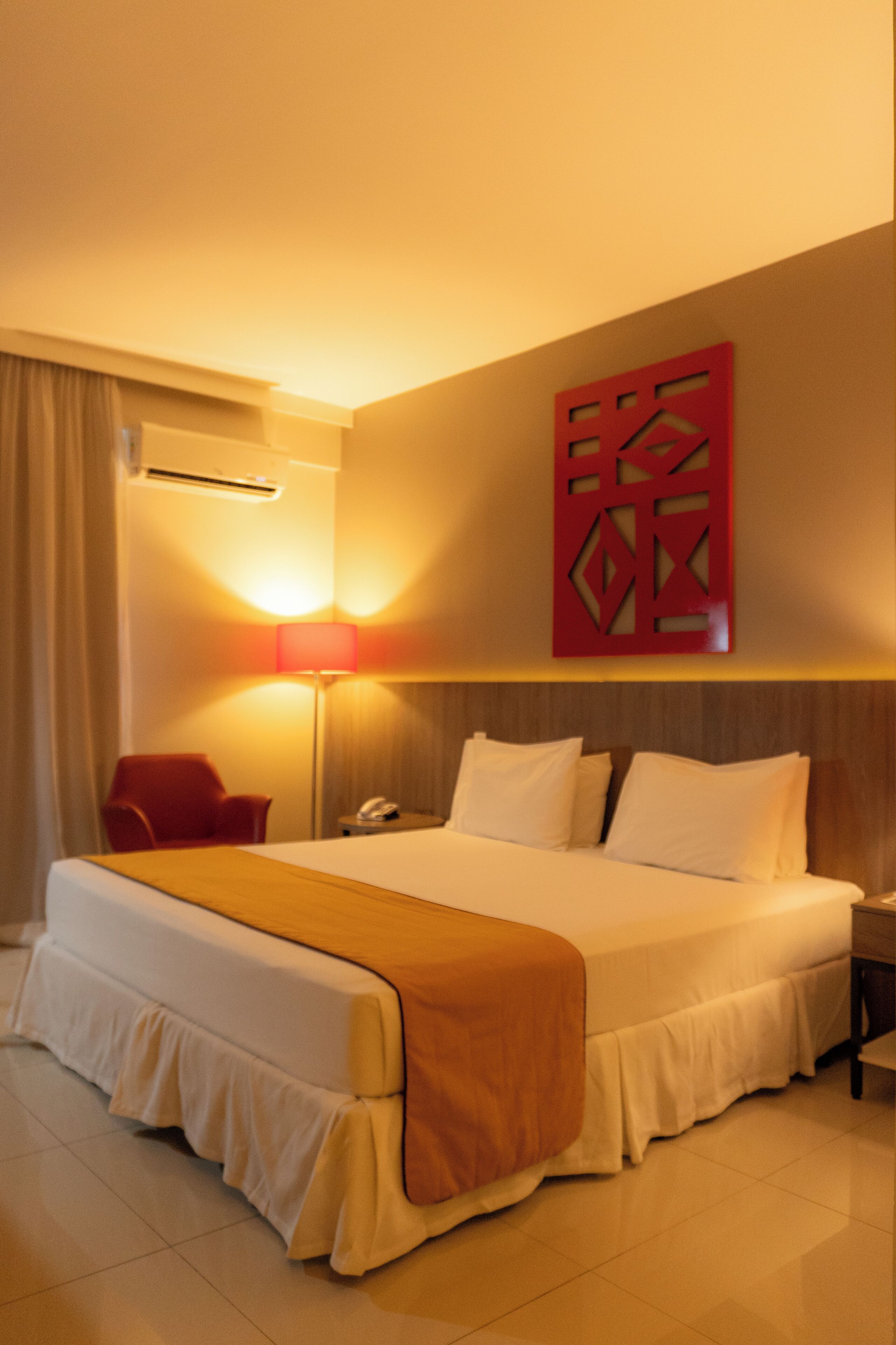 A hotel room with a large bed, a bedside table with a lamp, a red decorative wall piece, and a red chair.