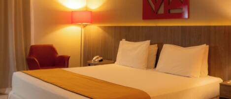 A hotel room with a large bed, a bedside table with a lamp, a red decorative wall piece, and a red chair.