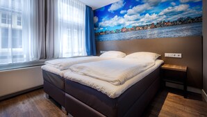 A bedroom with a large bed, a window with sheer curtains, and a mural of a coastal town on the wall. - Hotel Old Nickel (Amsterdam)