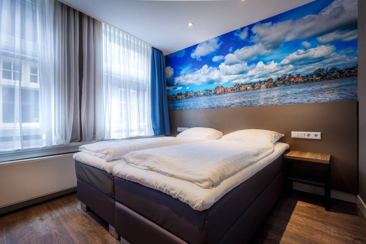 A bedroom with a large bed, a window with sheer curtains, and a mural of a coastal town on the wall.