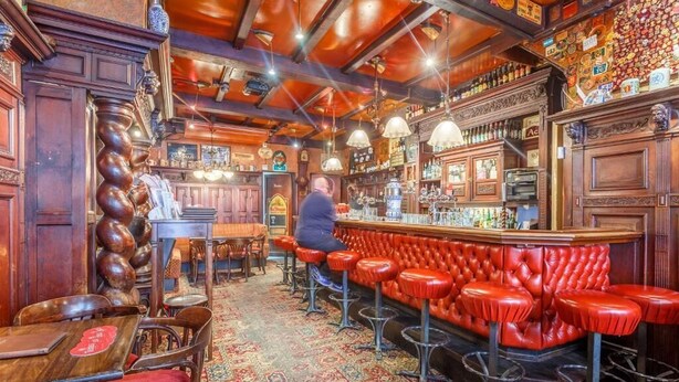 A bar with red leather stools, wooden bar counter, and a patterned carpet. - Hotel Old Nickel (Amsterdam)