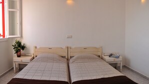 A double room with two beds, a bedside table with a lamp, a window with red curtains, and a bowl of fruit on the table. - Loukas Pension (Mykonos)