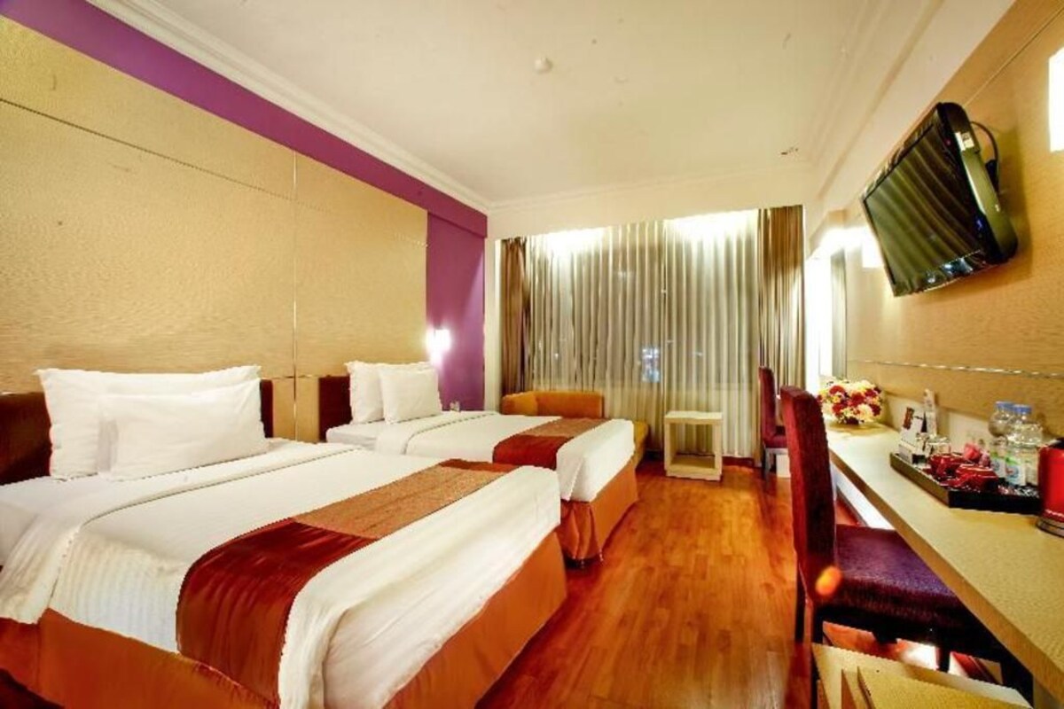 Ein Hotelzimmer mit zwei Betten, einem Schreibtisch, einem an der Wand befestigten Fernseher und einem Fenster mit Vorhängen.