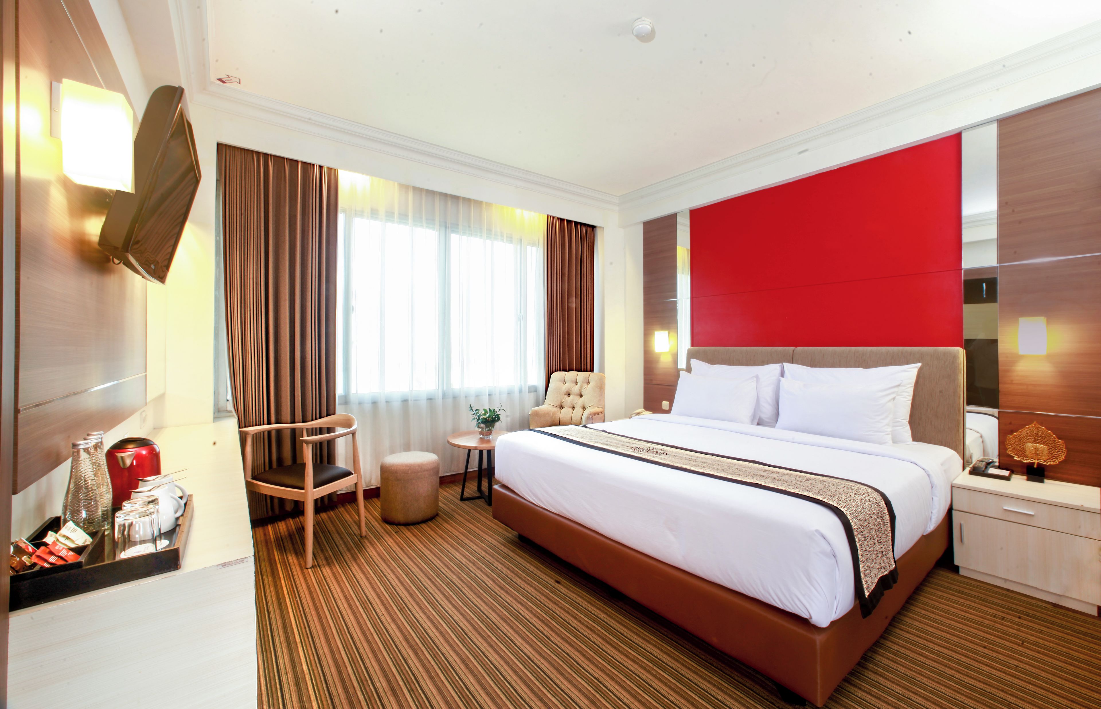 Una habitación de hotel con una cama grande, una mesita de noche de madera, una silla, una pequeña mesa con una planta y una pared de acento roja.