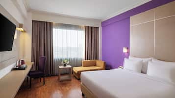 Ein Hotelzimmer mit einem großen Bett, einem Schreibtisch mit Stuhl, einem kleinen Tisch mit einer Vase und einem an der Wand befestigten Fernseher.