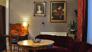 Una habitación con papel tapiz estampado, una mesa redonda con un tazón, un sofá de terciopelo rojo, una silla de madera y una lámpara.