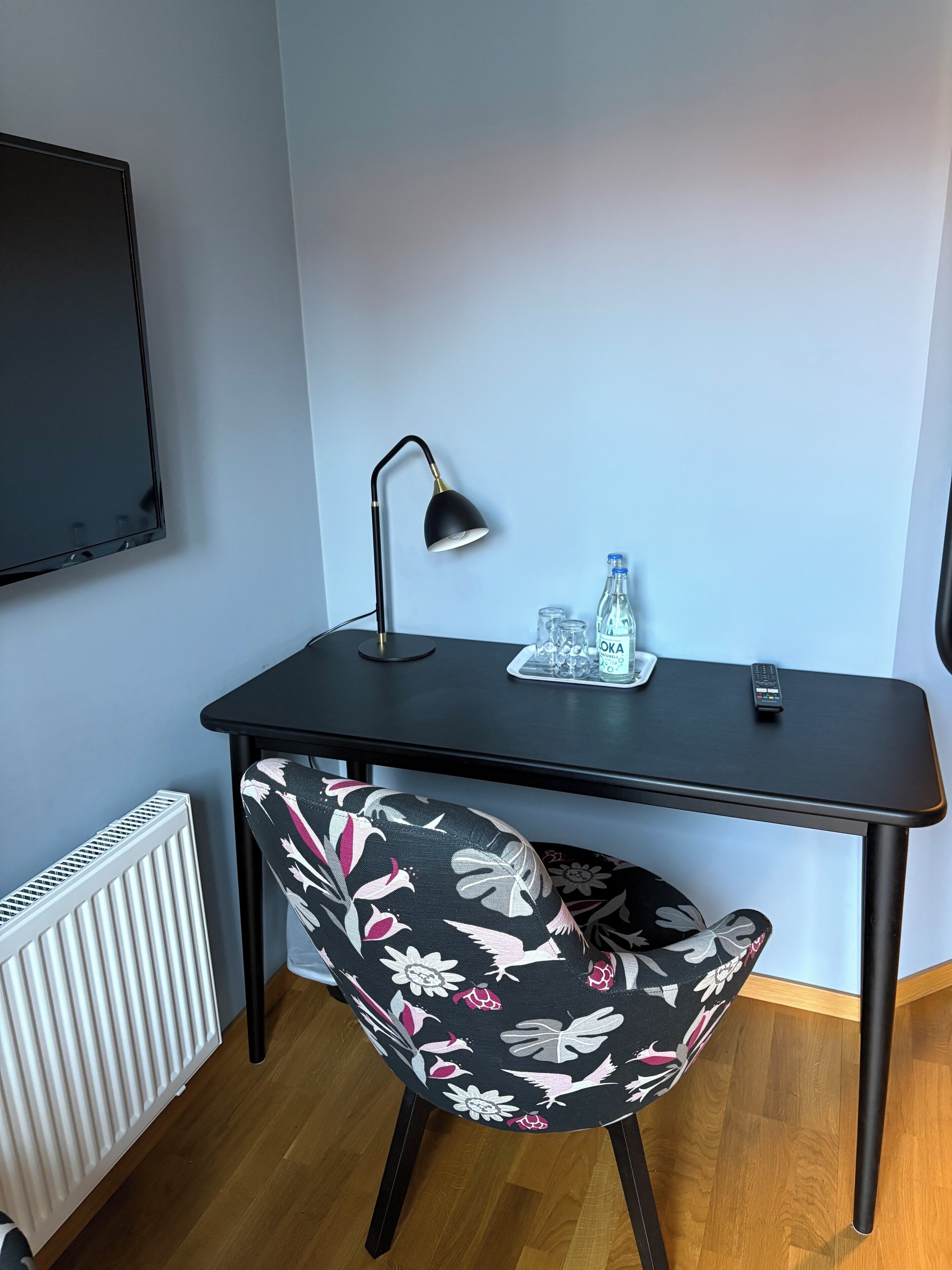 A small, modern room with a black desk, a black chair with a floral pattern, a black desk lamp, a remote control, and a bottle of water on the desk.